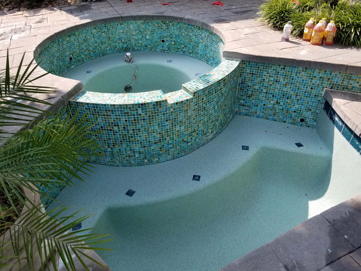 Another picture showing a pool and hot tub that have been drained of water for cleaning and remodeling work. A different view shows the pool surface is a light blue color and tiles around the pool and hot tub are a mixture of blue and green colors.