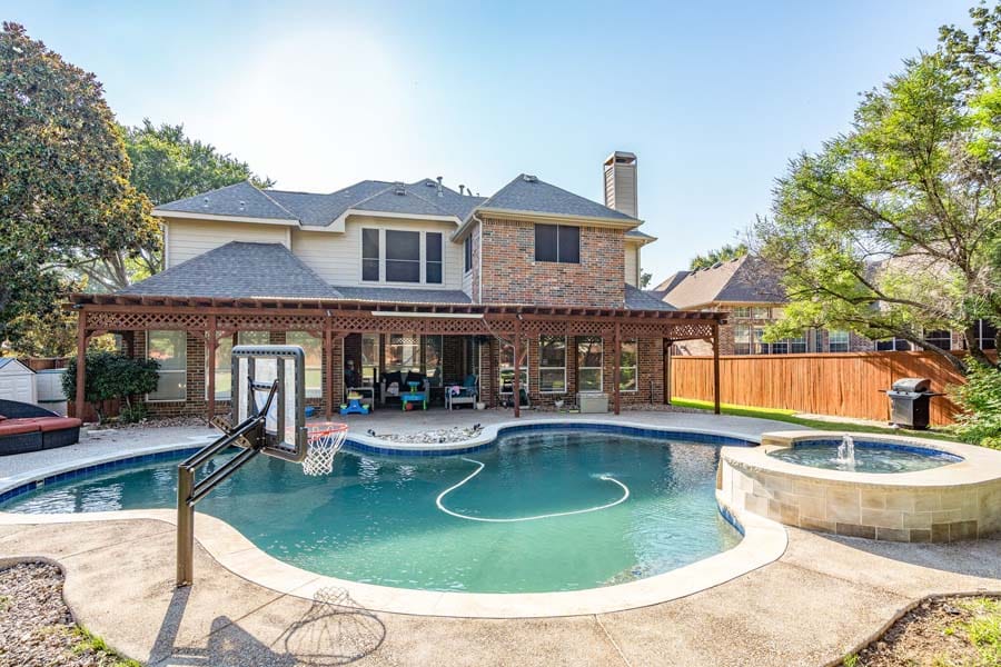 Weekly Pool Cleaning Service from Clear Choice Pool Care. Image taken from back of house, with sun shade, and table and chairs. House has brick facade. More lounging chairs and stools in front of windows